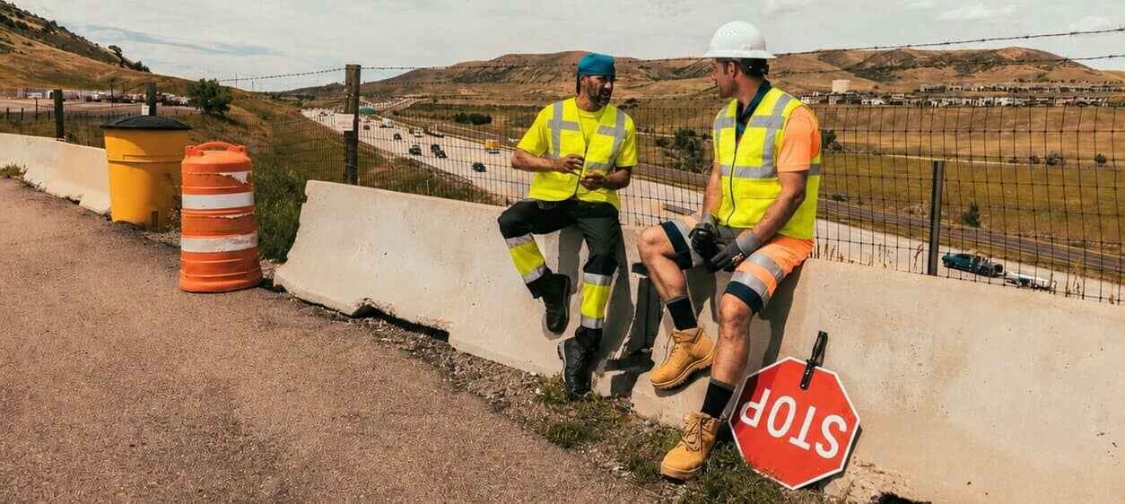 Two workers sit on a concrete barrier at the side of the road. They are wearing short-sleeved high-visibility clothing and cooling vests. In the background, you can see a barren mountain landscape and a cloudy, blue sky.