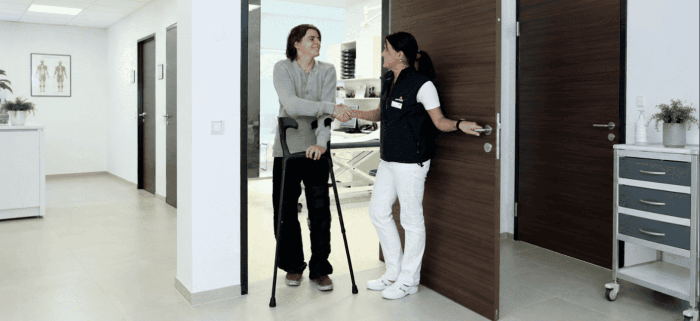 Two people are seen in the corridor of a clinic. A man with crutches shakes hands with a female doctor.