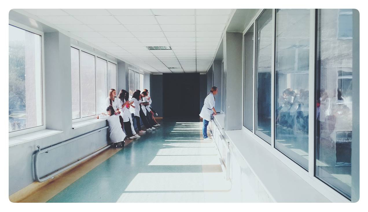 A corridor with windows on the right and left can be seen, where a group of medical staff are waiting while leaning against the wall.