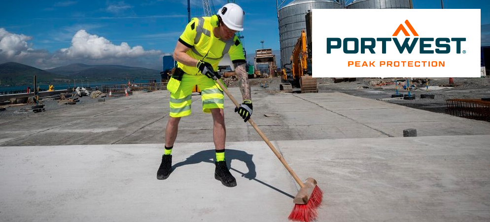A man in high-visibility yellow workwear and a white hard hat is sweeping the concrete floor in front of a factory. In the background, you can see mountains and a blue sky with small clouds. The blue and orange logo of the manufacturer, Portwest, is positioned in the upper right corner.