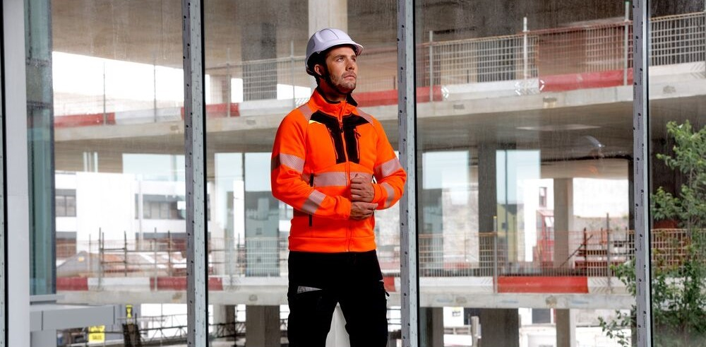 A worker wearing the DX4 high-visibility sweatshirt DX477. He is wearing a white hard hat and black trousers. In the background, several levels of a house under construction can be seen through a four-part window.