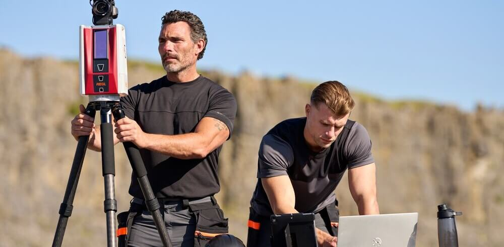 Two men in short-sleeved, black work clothes are working outdoors. One is taking measurements, the other is recording the results on a laptop. In the background, you can see rugged rocks and a blue sky.