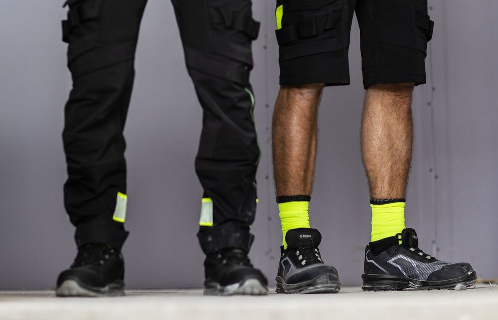 Two pairs of men's legs are shown: one man is wearing long black work trousers, the other is wearing short hi-vis yellow work shorts. A link to our work trousers category is included.