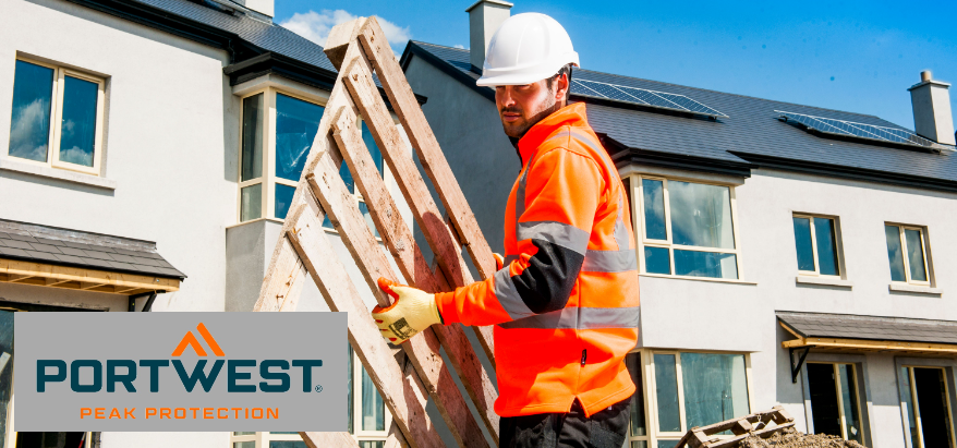 Worker wearing a white safety helmet, orange gloves, black trousers and a high-visibility orange sweater with reflective stripes. In the background you can see an almost completely built row of houses with white-painted walls and dark roofs. The sky is blue, some white clouds pass by.