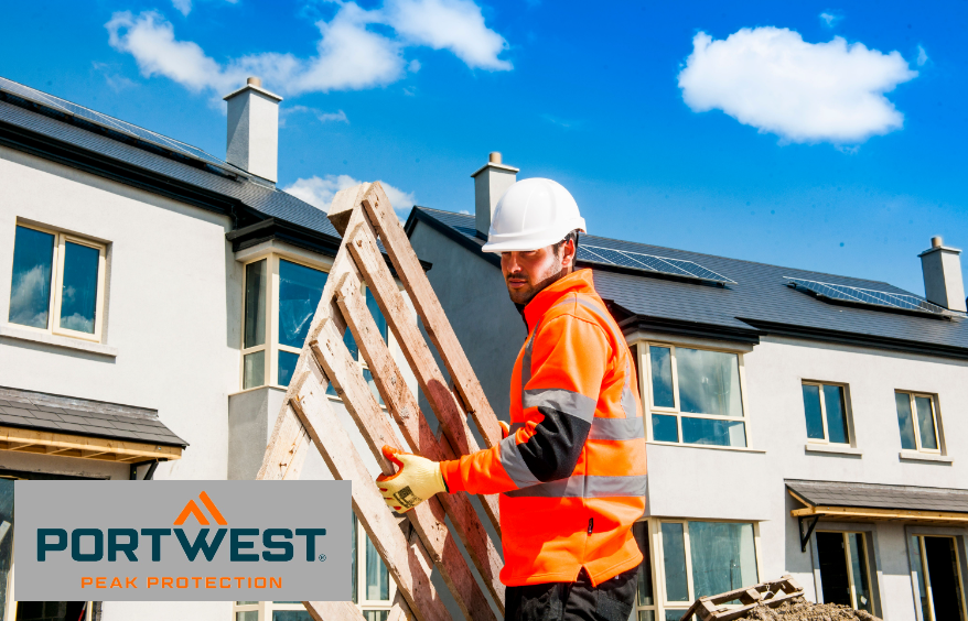 Worker wearing a white safety helmet, orange gloves, black trousers and a high-visibility orange sweater with reflective stripes. In the background you can see an almost completely built row of houses with white-painted walls and dark roofs. The sky is blue, some white clouds pass by. There is a link to our work sweaters.