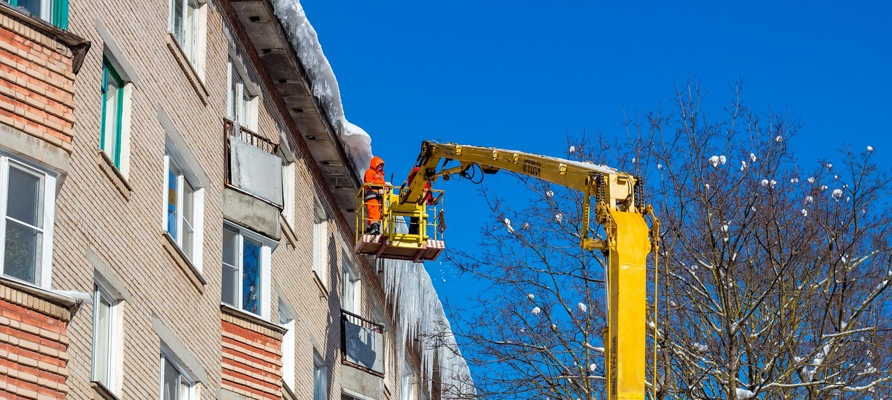 A row of houses with icicles on the roof ridge and snow on the roof. A worker in orange high-visibility clothing stands on a scissor lift. To the right, a bare tree with snow on its branches is visible.