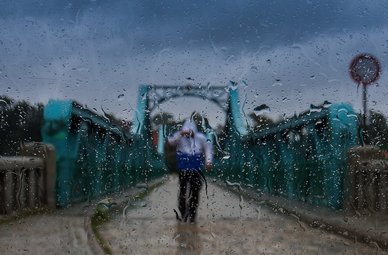 Blue bridge against a gray sky with a person in rain gear in the middle of the road. The scene was photographed through a wet pane of glass. A link to our entire selection of practical rainwear is provided.