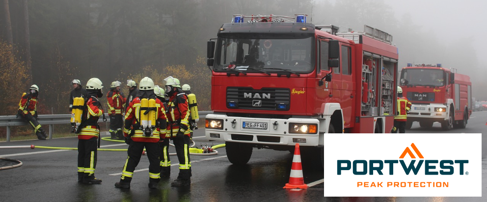 Street scene with two red fire engines and various firefighters. In the background you can see trees covered in fog. In the bottom right corner there is the blue-orange Portwest logo in front of a white rectangle.