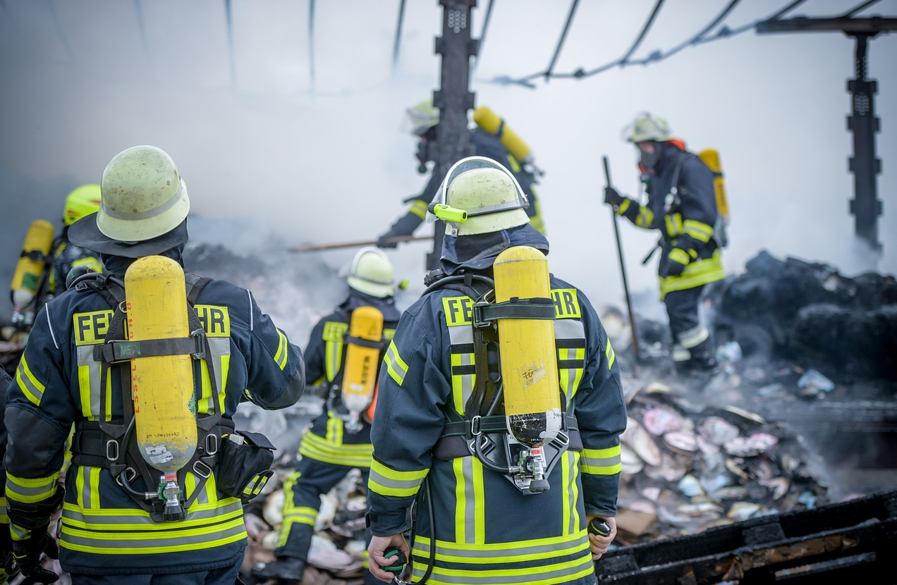 Firefighters in blue clothing with yellow warning stripes and helmets in a scrapyard. The link provided leads to our selection of flame-retardant firefighting clothing.