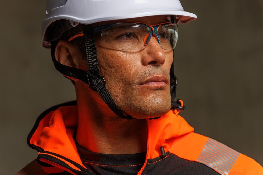 Close-up of a worker wearing a white safety helmet and clear protective goggles, looking into the distance with focus; dressed in high-visibility orange and black protective clothing. Links to our eye protection collection.