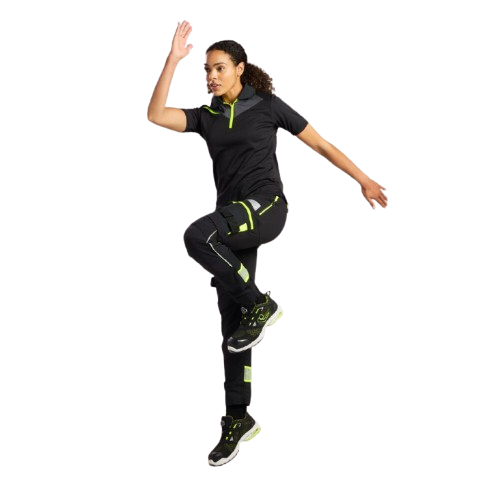Woman wearing black workwear with neon green details. She is moving her arms dynamically and is in the moment of jumping. A link to our women's workwear is included.