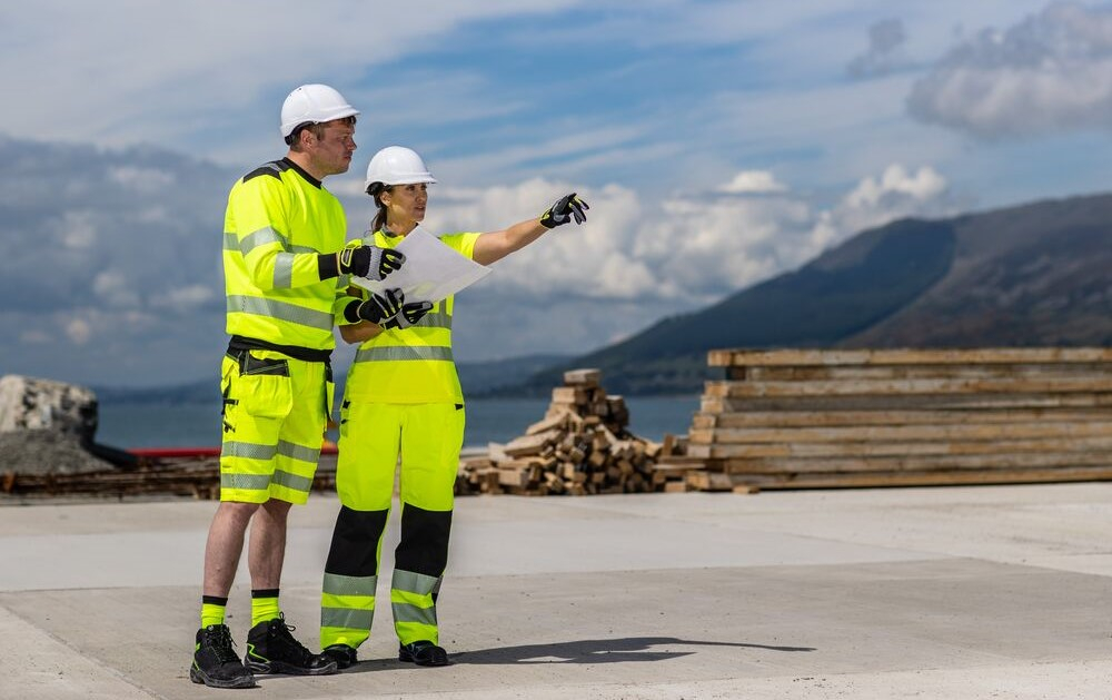 The image shows a man and a woman wearing yellow workwear and white safety helmets. They are standing on an asphalt surface and looking at a sheet of paper together. The woman is pointing at something with her left arm. In the background you can see mountains, a lake and a blue, slightly cloudy sky. A link to our women's workwear is included.