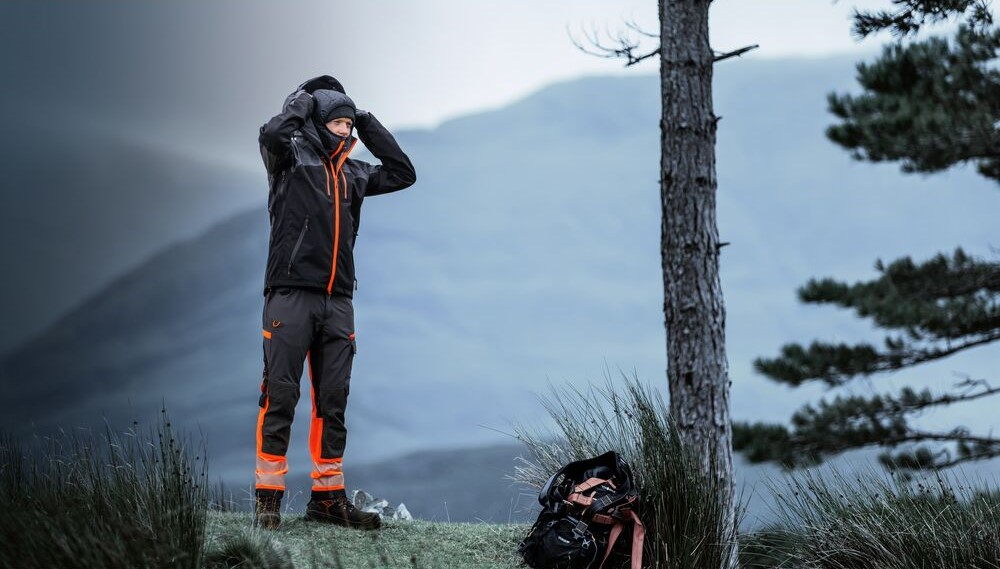 Worker in weatherproof workwear with reflective elements is outdoors in the mountains putting on a winter beanie. A link to our beanies is included.