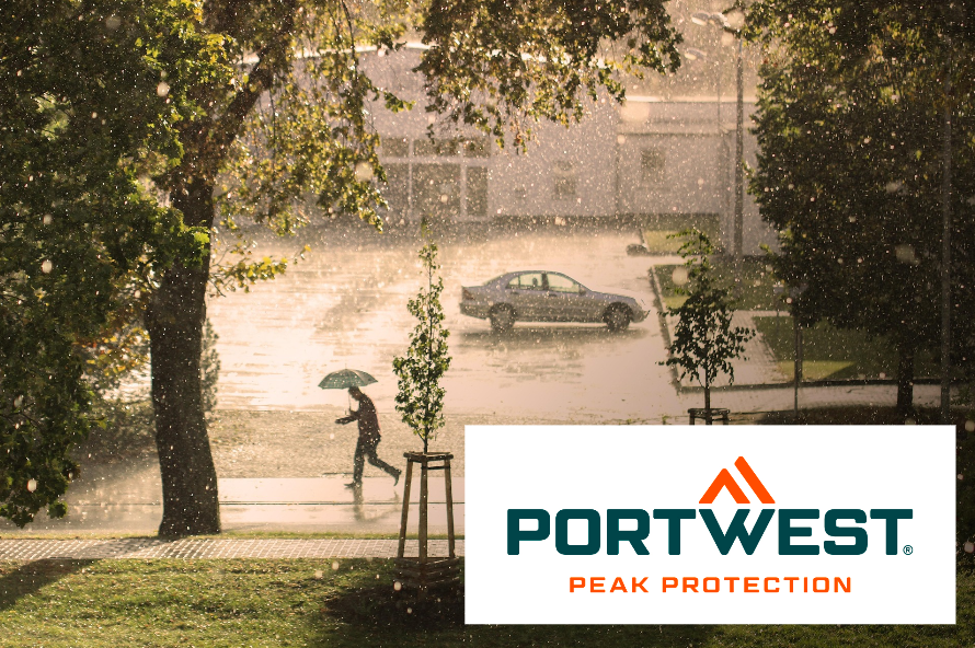 View of a street in the rain. A person with an umbrella crosses the street and the sun is shining even though it is raining. The logo of the manufacturer Portwest is in the bottom right of the picture. A link to our rainwear is provided.