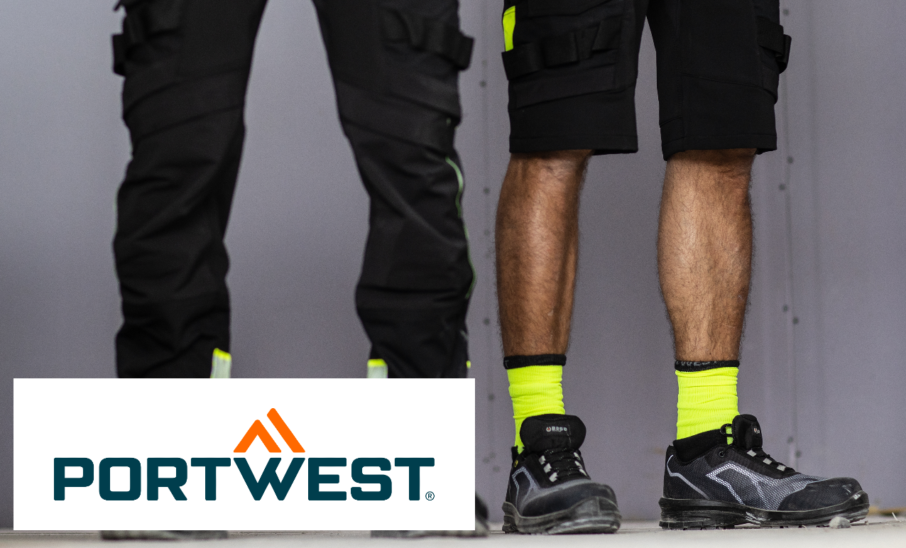 Image of two pairs of legs: one person wears long black work trousers, the other wears black work shorts, neon yellow socks and black work shoes. The floor is light gray, the wall in the background is darker gray. There is a link to our entire collection of short work trousers.