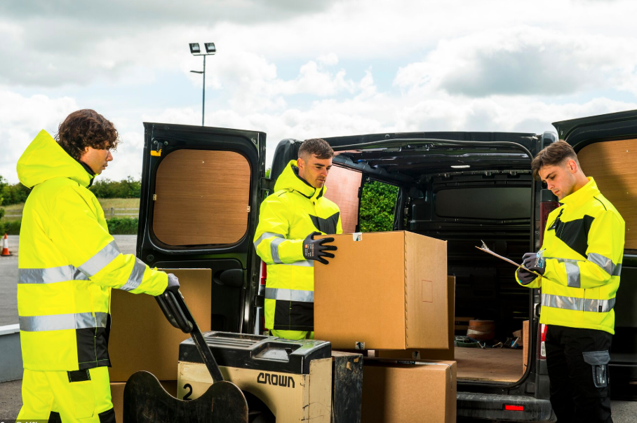 Three men in high-visibility yellow softshell jackets with reflective stripes work in front of an open van. One is pushing a pallet of boxes with a pallet truck, another is lifting a large box from the van, and the third is writing on a clipboard. The scene takes place outdoors under a cloudy sky. There is a link to our softshell jackets.