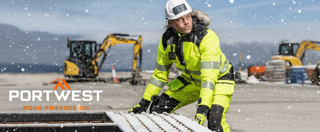 A worker in yellow, reflective winter protective clothing works outdoors in snowfall. He wears a hard hat and gloves. Construction vehicles and a snowy landscape can be seen in the background. In the lower left corner, the logo of the manufacturer Portwest is in white and orange lettering.