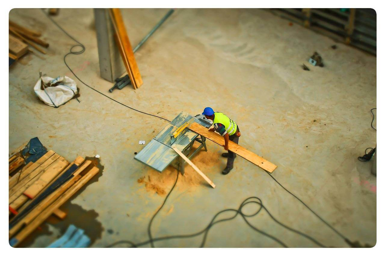 A worker in a yellow high-visibility vest is seen from a bird's-eye view. He is sawing a wooden board on a table saw.