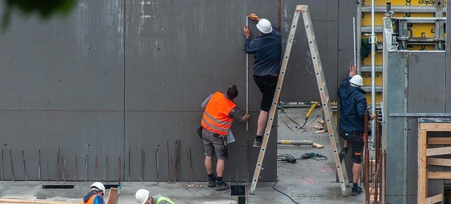 Two workers are seen on a construction site. One is standing on a ladder, the other is holding it. Both are wearing work shorts; one is wearing a white helmet and the other an orange high-visibility vest.