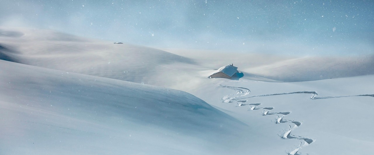 A snowy mountain scene with a solitary alpine pasture. Ski tracks in the snow lead to the cabin. The sky is bright blue, and snowflakes gently fall across the landscape.