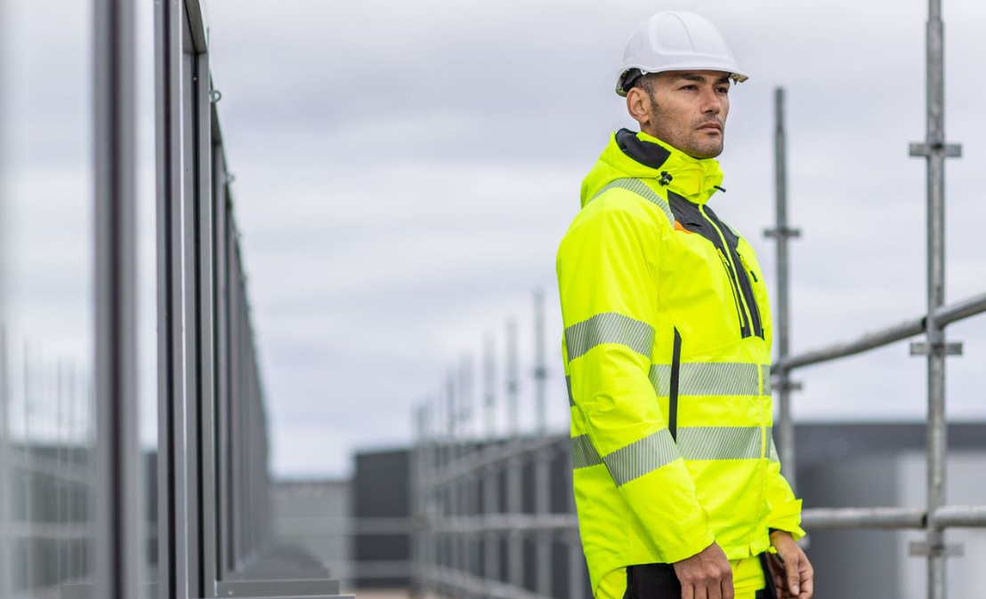 A worker in high-visibility yellow work clothing with a white helmet in front of a window front and the upper part of a scaffolding with a link to the DX4 collection.