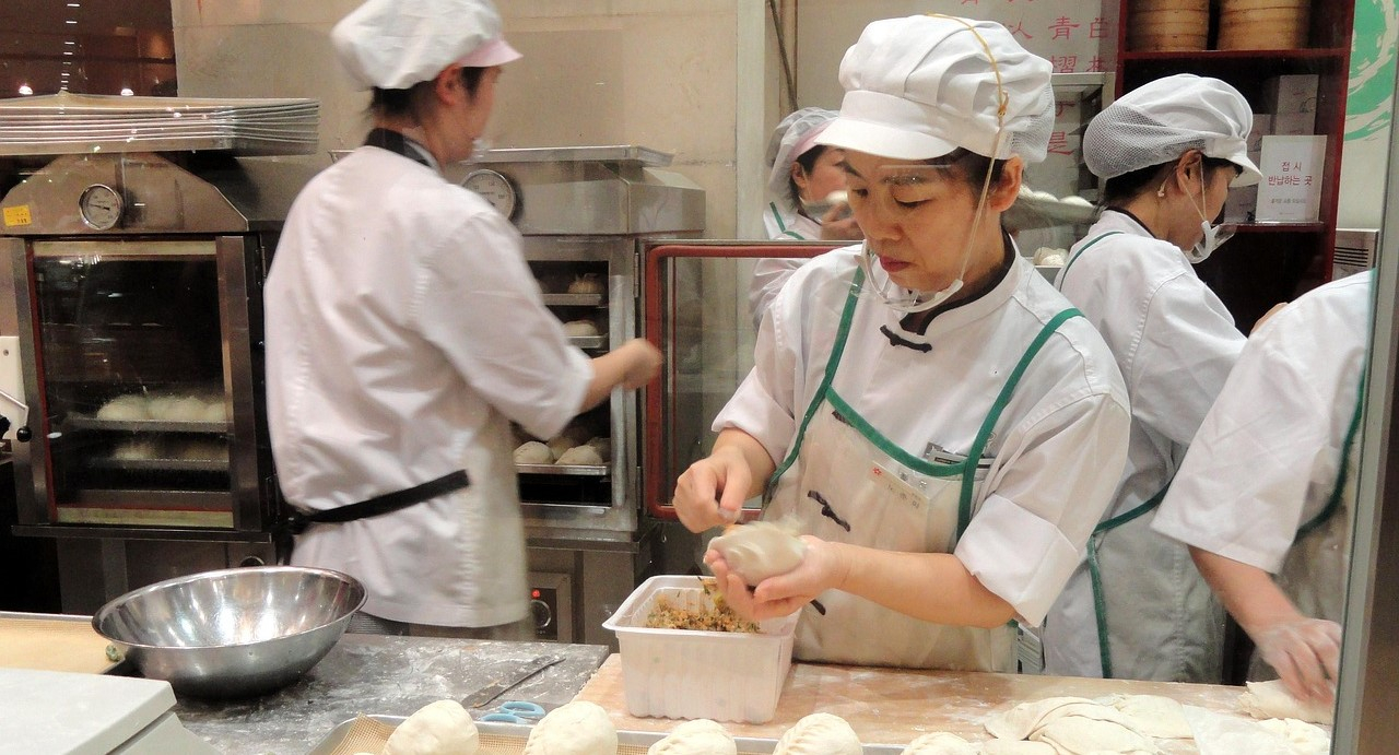 A hectic kitchen scene with five female chefs in white chef clothing, including aprons, hats, and face masks. The chef in the foreground is filling dumplings, while others operate the oven and carry trays of baked goods.