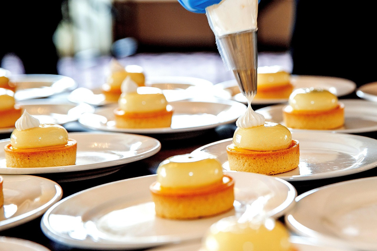 Plate with fine pastry. A hand wearing a blue glove holds a piping bag and applies a white mixture onto a small cake.