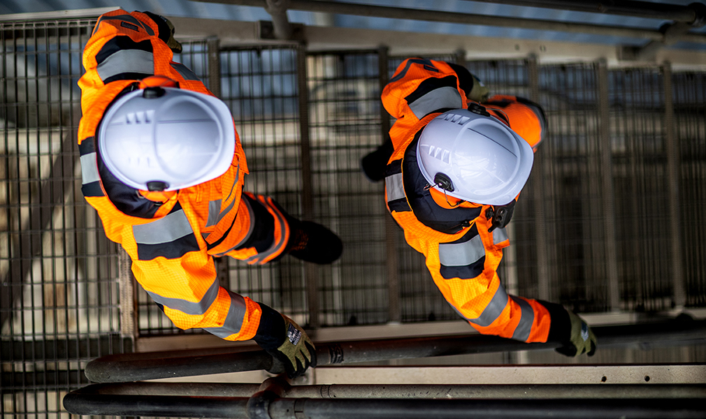 Bird's eye view of two workers wearing white hard hats, black and green gloves, safety boots and orange hi-vis coveralls with link to our Arc Flash protective clothing collection.