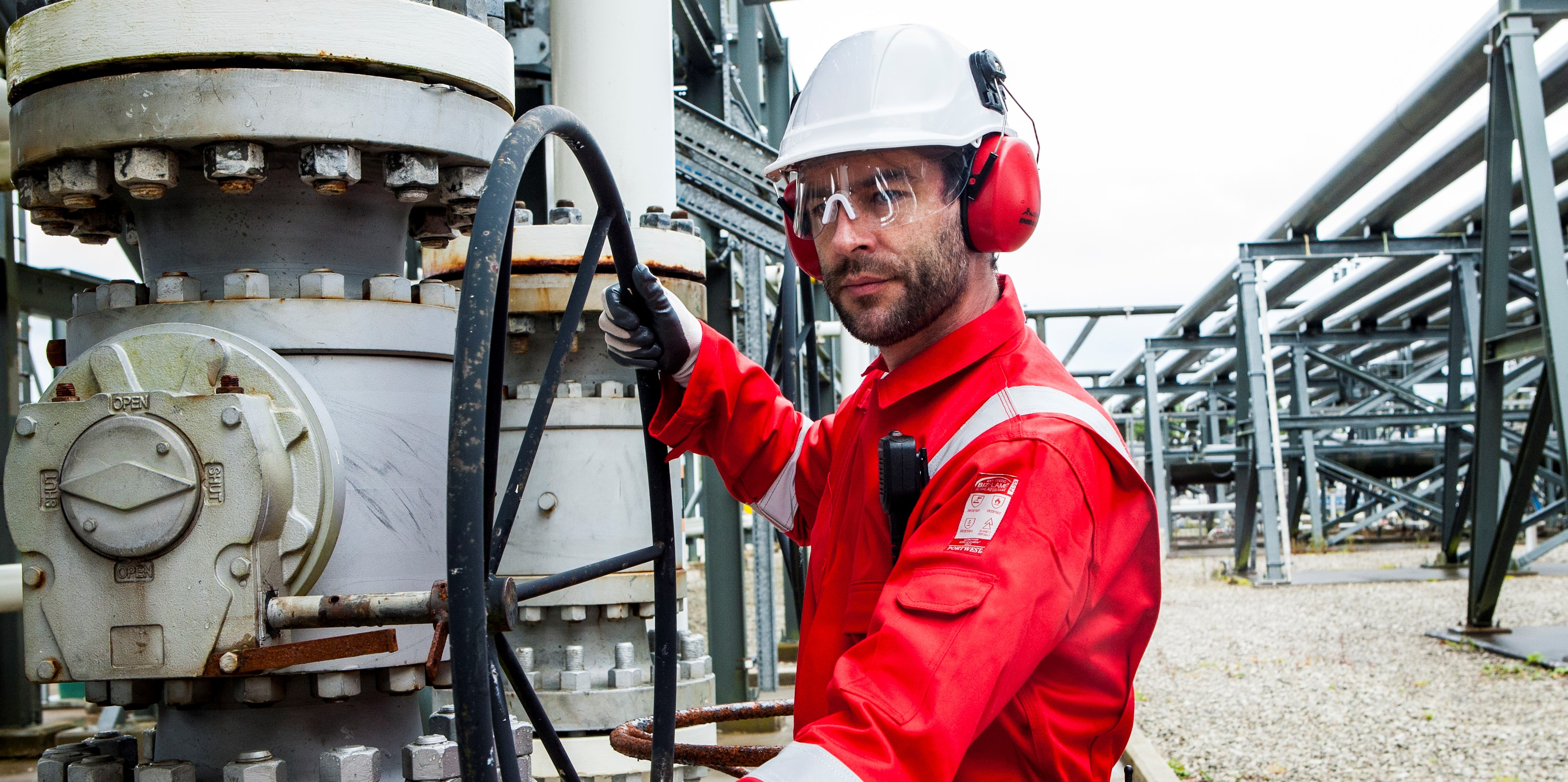 Worker in an industrial context wearing protective clothing: white helmet, goggles, red earmuffs, black gloves and red FR50 overalls with reflective strips.