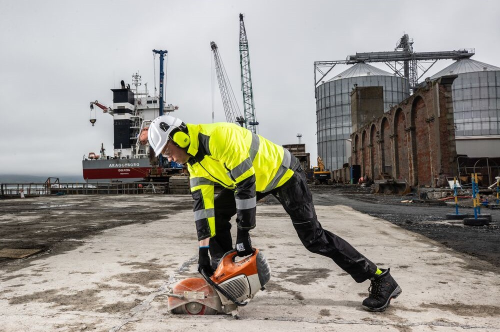 A worker wearing a yellow high-visibility jacket and black work trousers cutting a base plate with a circular saw. Large silos and a container ship can be seen in the background. The weather is grey and rainy.