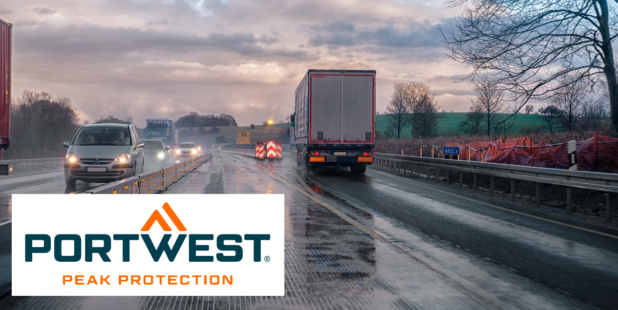 Highway with cars and trucks driving on wet pavement with lights on. The sky is blue and crossed by gray clouds; on the right side of the picture you can see a lush green meadow and bare trees. In the lower left edge of the picture is the orange-blue Portwest logo on a white, rectangular background.