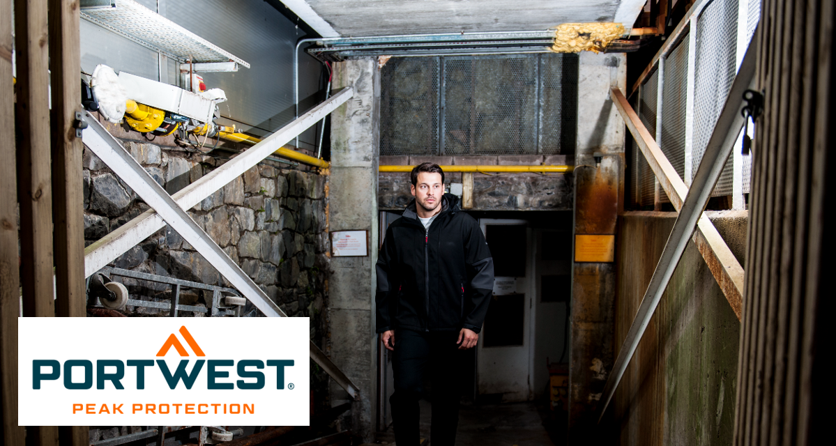 Looking into a stable with yellow pipes on the wall and a worker in the middle. The man is wearing the waterproof softshell jacket with hood TK53 in black and gray. In the lower left edge of the picture is the orange-blue Portwest logo on a white, rectangular background. There is a link to our collection of softshell jackets.