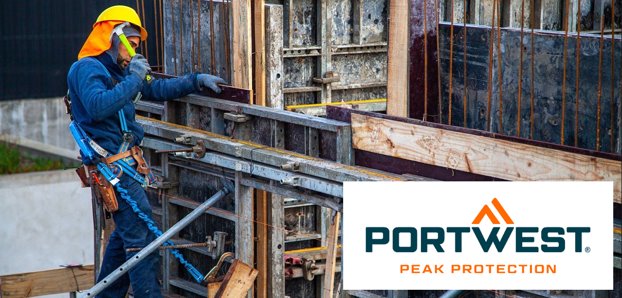 Workers in blue work clothes with a yellow safety helmet on a scaffolding in front of the shell of a house. He is secured with a height safety device and holds a hammer in his right hand. The Portwest logo is in the lower right area of the image.