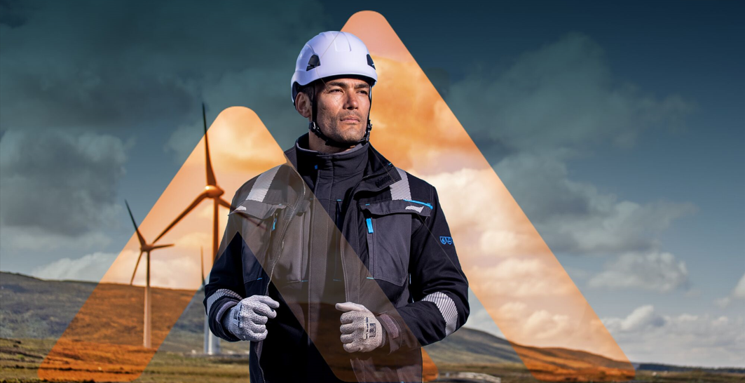 A man wearing a safety helmet, gloves, and a work jacket stands in front of wind turbines; illustrating safety at work and fall protection for working at heights.