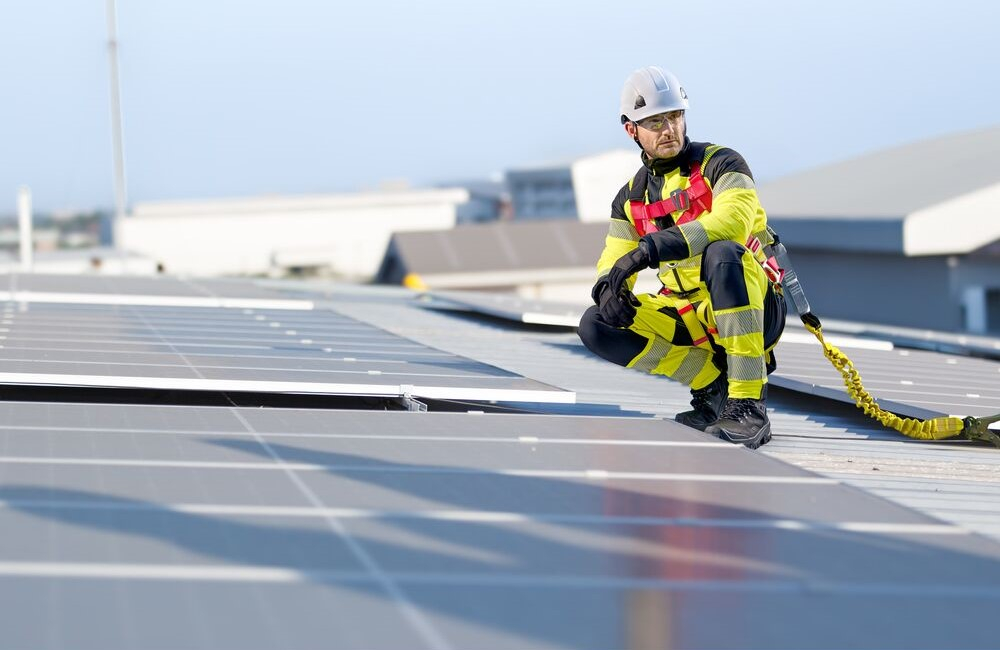 Worker on a roof among photovoltaic panels. Wearing high-visibility yellow workwear, a white helmet, black gloves and shoes. Secured with Portwest 3-Point Plus FP18 harness. Link to our fall protection.