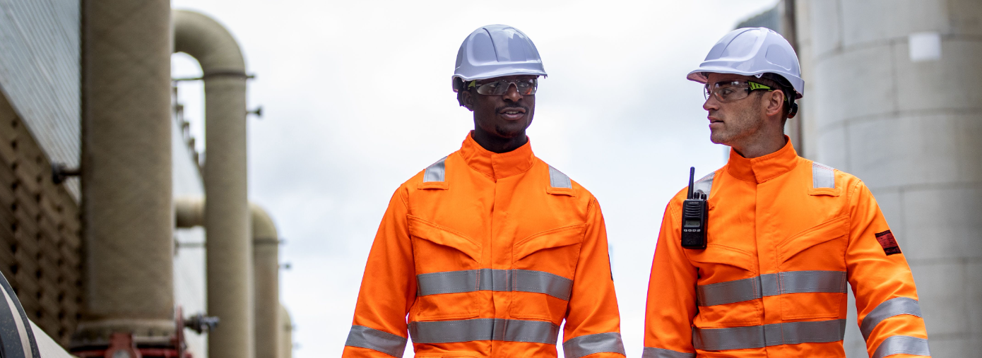 Two workers in front of industrial background wearing orange high visibility clothing and white hard hats.