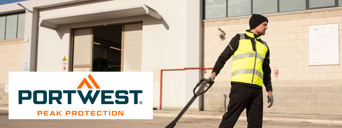 A worker in black work clothes with a high-vis yellow vest in front of a warehouse with the gate open. The worker pulls a pallet truck. In the bottom left of the picture is the orange and blue Portwest logo against a white background.