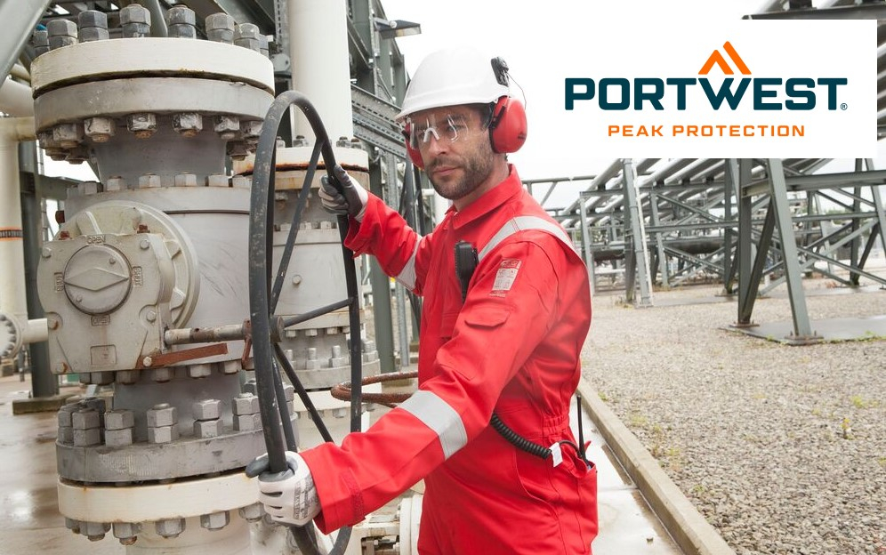 An industrial worker is wearing a red protective suit with reflective stripes, a white hard hat, hearing protection, and safety goggles. He is operating a large black handwheel on an industrial plant, presumably in a refinery or industrial yard. Pipelines and steel structures can be seen in the background. The Portwest brand logo with the slogan "Peak Protection" is located at the top right of the image. A link to our flame-retardant overalls is provided.