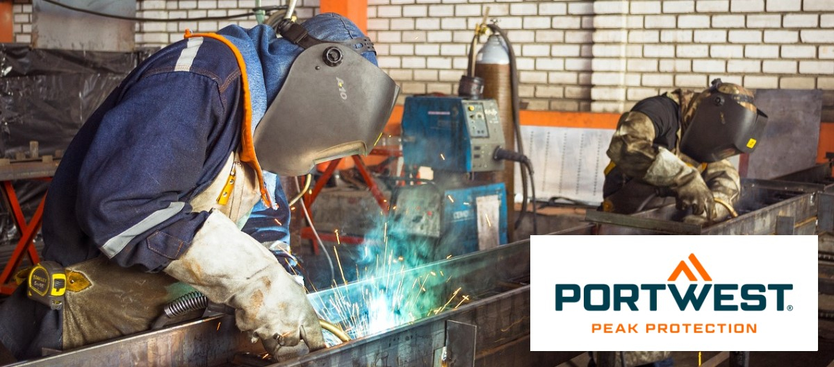A view into a welding shop: two workers in flame-resistant protective clothing, gloves, and welding masks are welding a long sheet of metal.