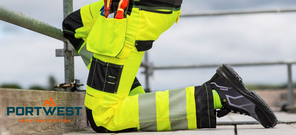A construction worker kneels on a scaffolding floor. He is wearing bright yellow high-visibility work trousers with integrated knee pads and black safety shoes. A cloudy sky can be seen in the background. The manufacturer's logo, Portwest, in blue and orange letters is in the lower left corner.