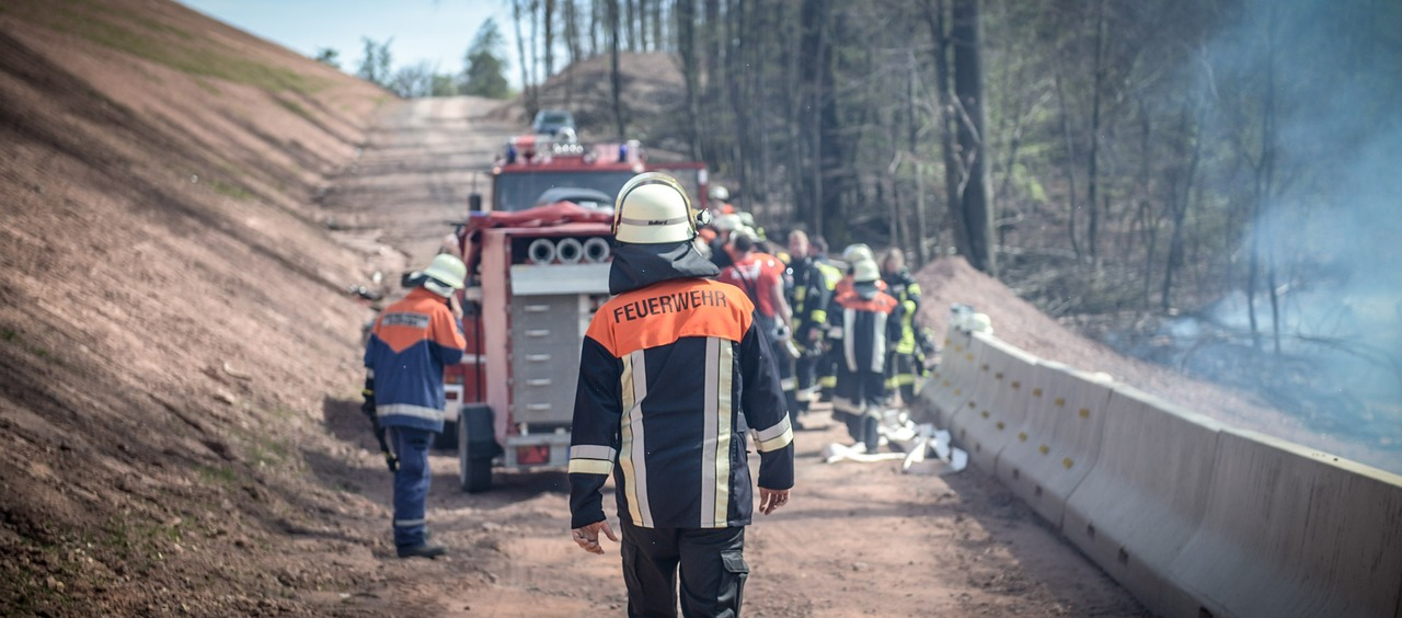 Rear view of a firefighter in full gear. In the background you can see a fire engine on a forest path and a group of firefighters at work. The link to the FR98 FR98 Bizflame FR98 FR98 Bizflame FR Fire Retardant Coverall is provided.