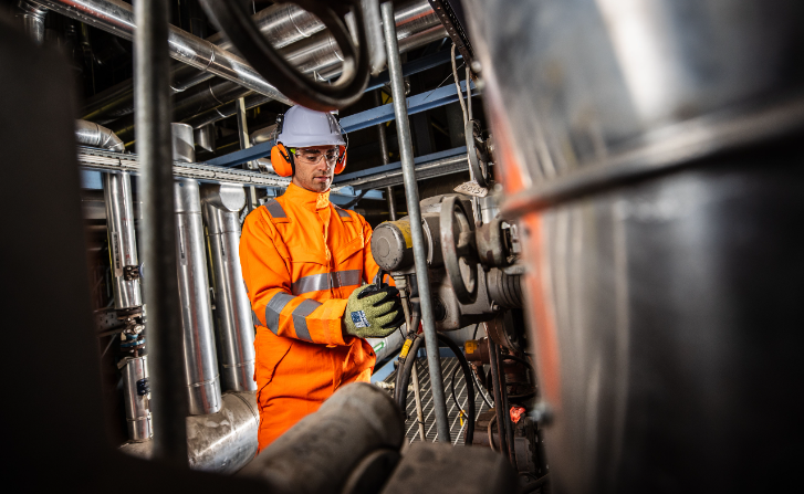 Worker with white helmet and ear protection in orange workwear with gloves against an industrial background. The image is linked to the collection of work gloves.