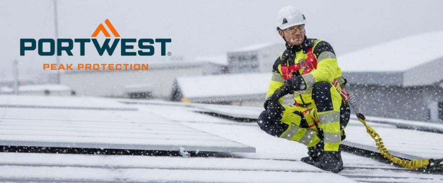 A worker in a yellow high-visibility coverall with reflective stripes kneels on a snow-covered roof next to a photovoltaic system. He is wearing a white hard hat and fall protection. More snow-covered roofs are visible in the background, and the manufacturer's logo, Portwest, is visible in blue and orange in the upper left of the image.