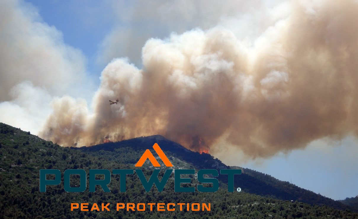 A wildfire rages on a green mountainside, thick clouds of smoke rising into the sky. A firefighting aircraft is approaching, dropping water or extinguishing agent over the flames. The Portwest brand logo is visible in the foreground – the slogan "Peak Protection" is below. A link to our selection of firefighting clothing is included.