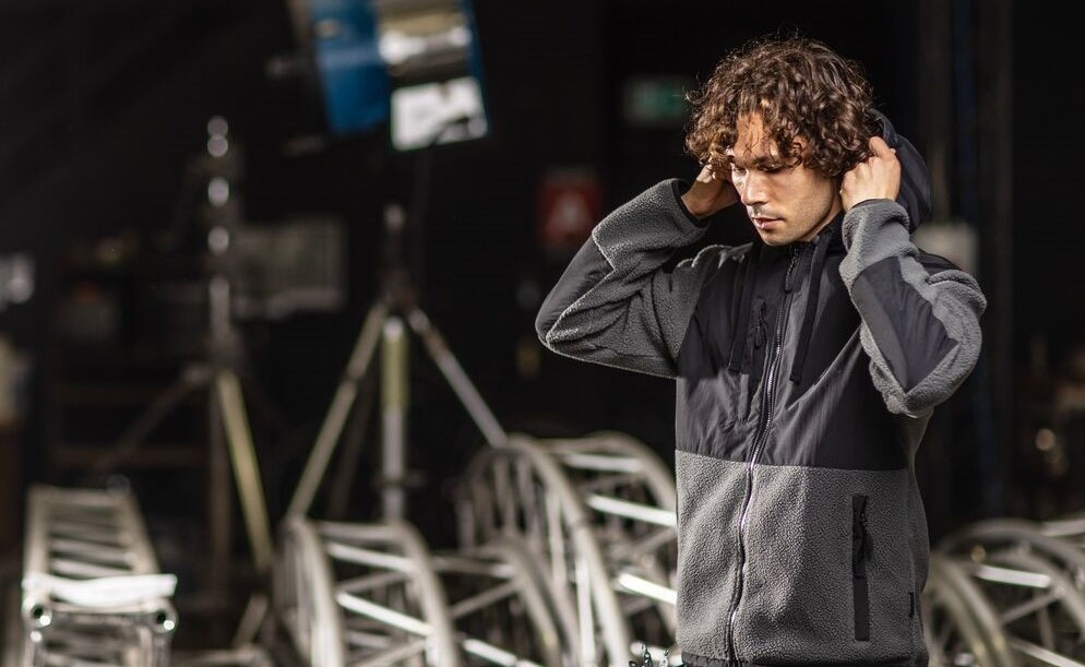 Man with curly hair wearing a grey fleece jacket with hood and black panels, standing in an industrial setting with trusses and spotlights. A link to our fleece collection is included.