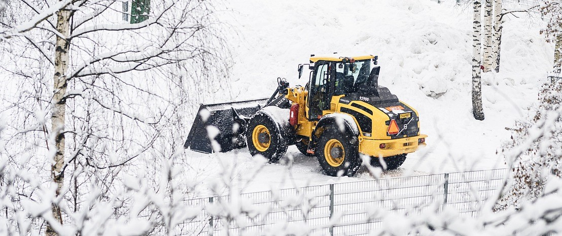 Yellow tractor with shovel in snowy landscape with fences and birch trees.