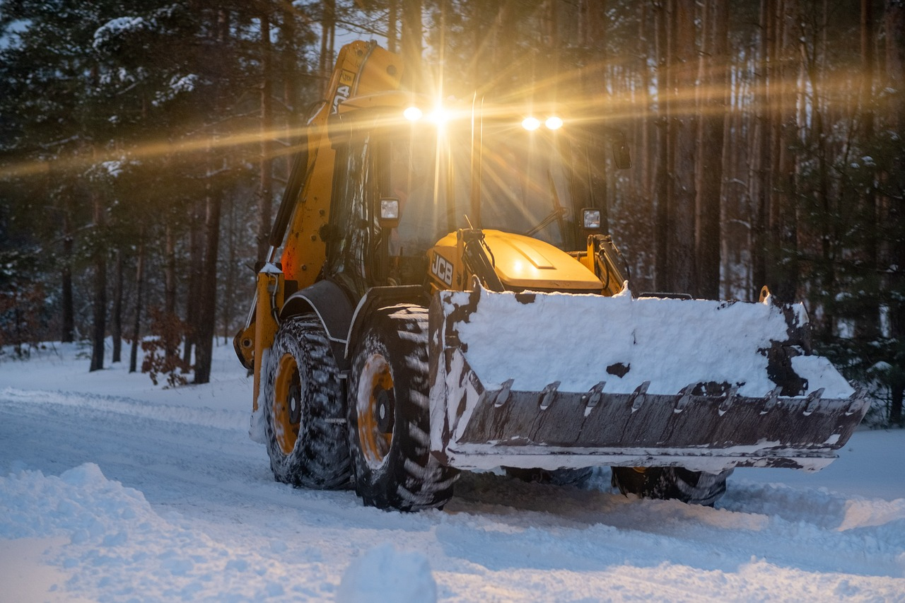 Yellow tractor with a large shovel on a snowy forest path. Twilight is slowly falling and the vehicle has a light on.